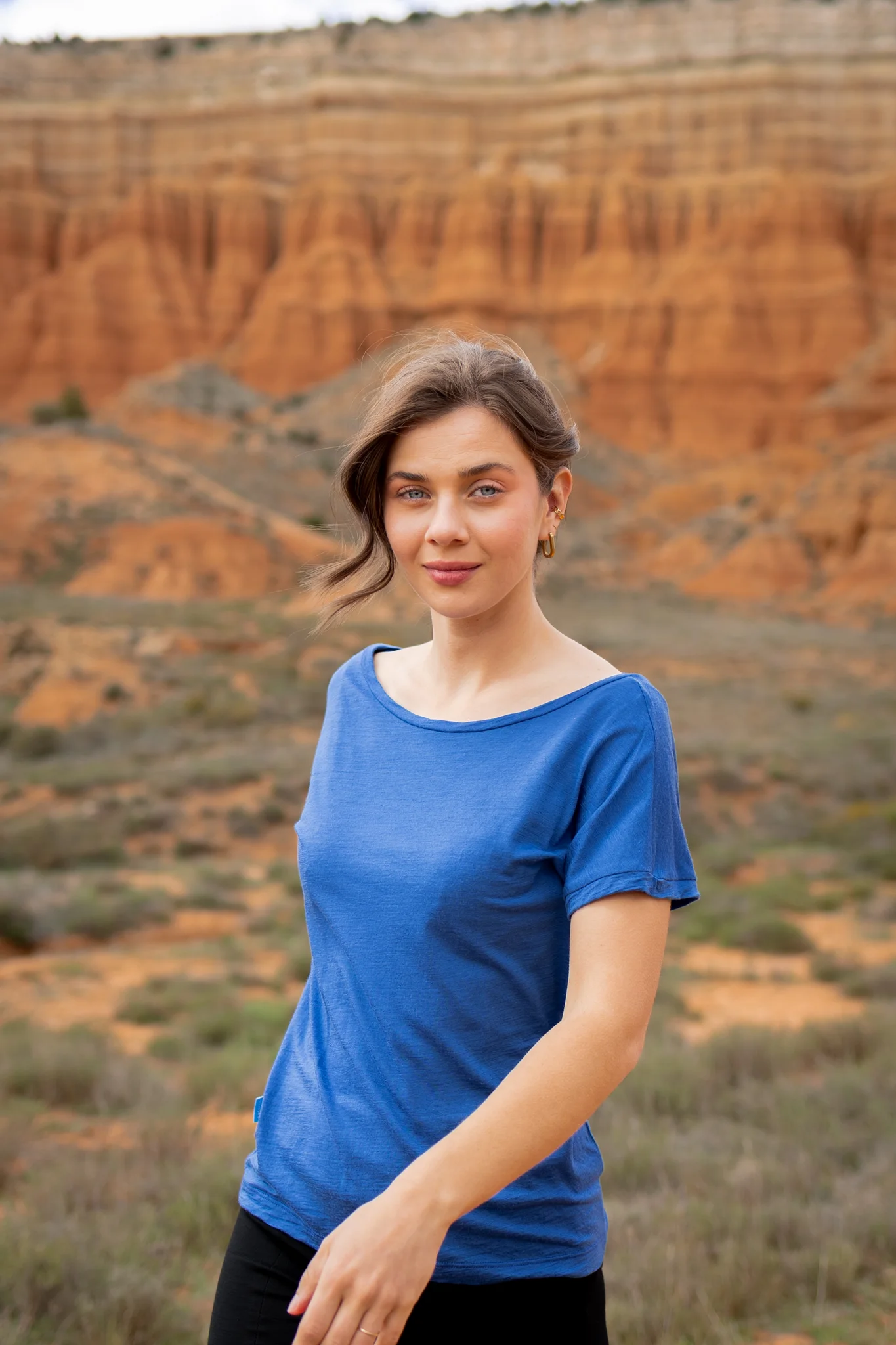 Kobieta o falowanych brązowych włosach nosi koszulkę z wełny merino prosta damska mokka, stojąc na zewnątrz wśród czerwonych formacji skalnych i rzadkiej roślinności, patrząc w kamerę z lekkim uśmiechem.