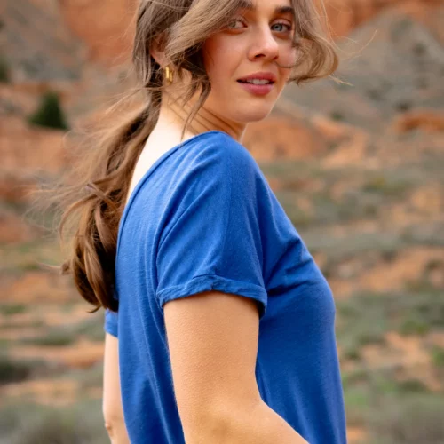 Kobieta z długimi brązowymi włosami spiętymi w kucyk nosi koszulkę z wełny merino prosta damska mokka, stojąc na zewnątrz w skalistym pustynnym krajobrazie. Spogląda przez ramię z lekkim uśmiechem, gdy wiatr porusza jej włosami.