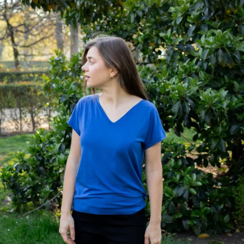 Kobieta o długich brązowych włosach, ubrana w koszulkę z wełny merino prosta damska mokka, stoi na trawie przed dużym zielonym krzewem liściastym, patrząc w lewo.
