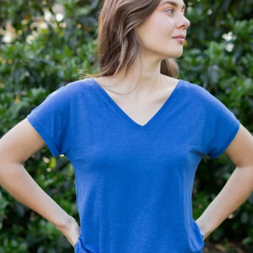 Kobieta z długimi brązowymi włosami stoi na zewnątrz przed zielonymi liśćmi, ubrana w koszulkę z wełny merino prosta damska mokka i czarne spodnie, ręce na biodrach, patrząc w bok.