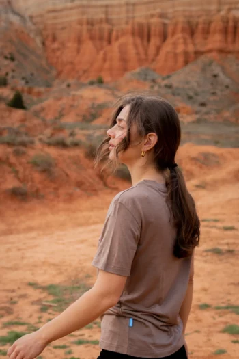 Kobieta z długimi brązowymi włosami spiętymi w kucyk nosi koszulkę z wełny merino prosta damska mokka, stojąc na zewnątrz z zamkniętymi oczami w czerwonym skalistym pustynnym krajobrazie.