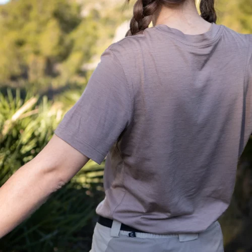 Osoba z brązowymi włosami zaplecionymi w warkocz, ubrana w koszulkę z wełny merino prosta damska mokka i beżowe spodnie, stoi na zewnątrz z lekko wyciągniętymi ramionami, otoczona zielenią i rozmytymi drzewami w tle.