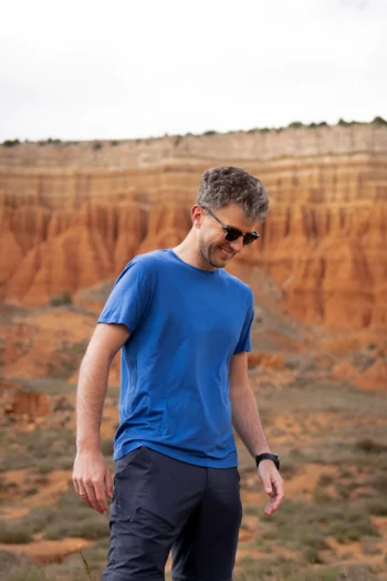 Kobieta ubrana w mokkę Koszulka z wełny merino prosta damska uśmiecha się na zewnątrz z czerwonymi formacjami skalnymi i warstwowymi klifami w tle.