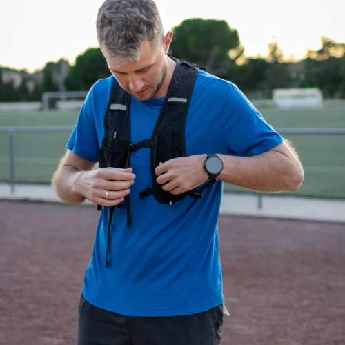 Kobieta w koszulce z wełny merino prosta damska mokka stoi na bieżni, dopasowując czarną kamizelkę do biegania. Ma krótkie włosy i nosi zegarek na lewym nadgarstku, a w tle widoczne jest boisko sportowe.