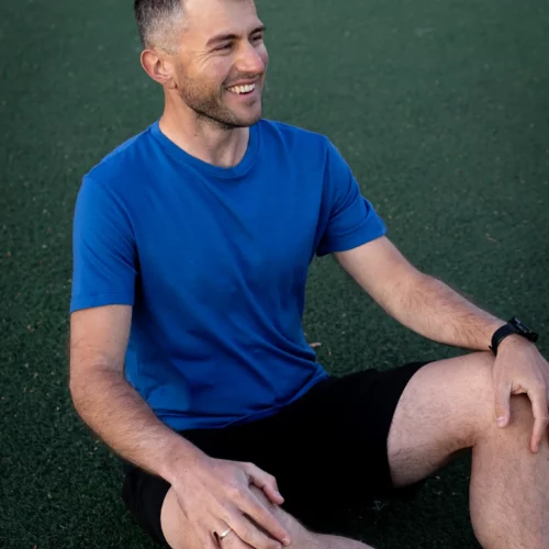 Kobieta ubrana w koszulkę z wełny merino prosta damska mokka siedzi na zielonej sztucznej murawie, uśmiechając się i patrząc w bok z jedną ręką na kolanie.