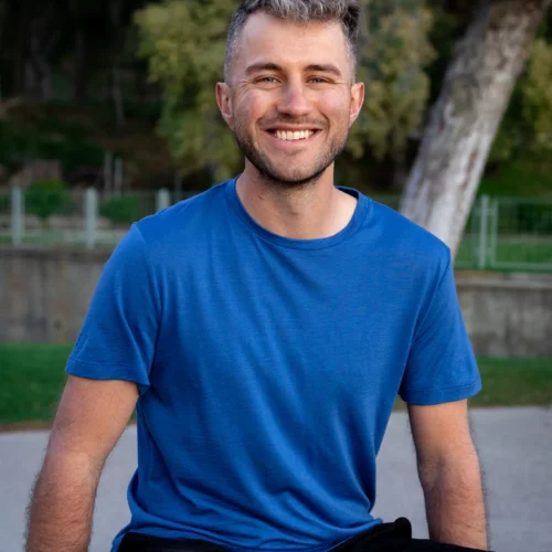 Mężczyzna z krótkimi jasnobrązowymi włosami i brodą siedzi na zewnątrz na betonie, uśmiechając się. Jest otoczony zielenią i ma na sobie Koszulkę z wełny merino prosta damska mokka.