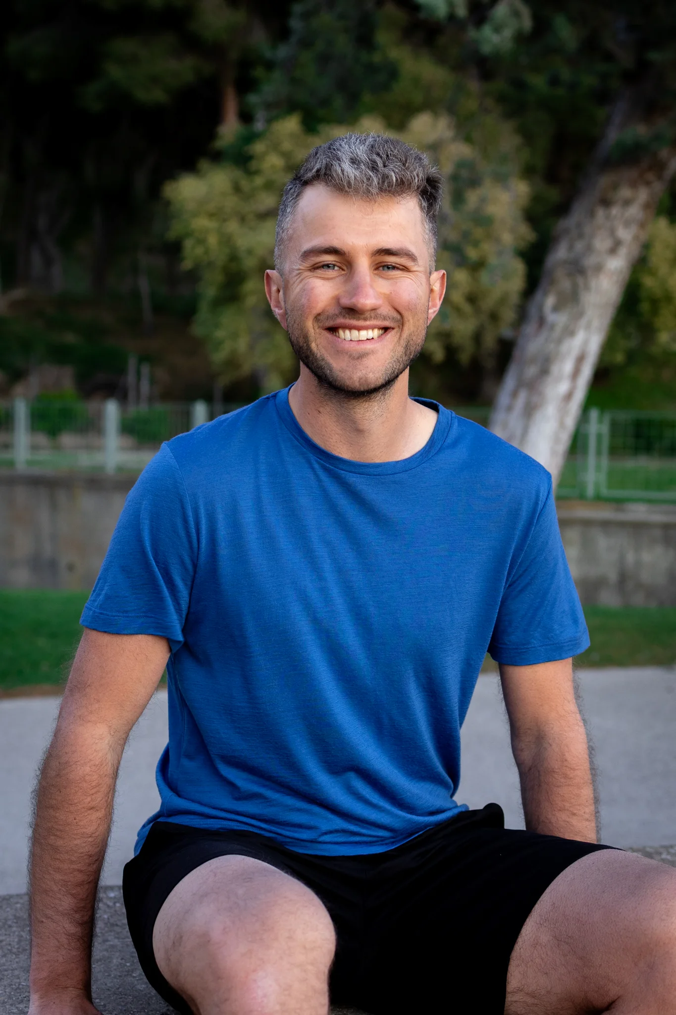 Mężczyzna z krótkimi jasnobrązowymi włosami i brodą siedzi na zewnątrz na betonie, uśmiechając się. Jest otoczony zielenią i ma na sobie Koszulkę z wełny merino prosta damska mokka.