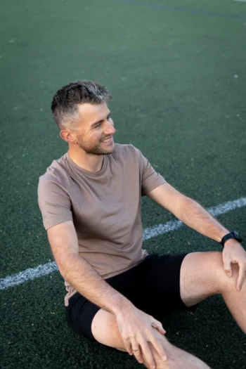 Kobieta w mokasynu Koszulka z wełny merino prosta damska siedzi na sztucznej trawie, uśmiecha się i rozgląda na boki. Ma krótkie włosy i nosi zegarek na lewym nadgarstku. Na ziemi pojawiają się białe linie boiska sportowego.