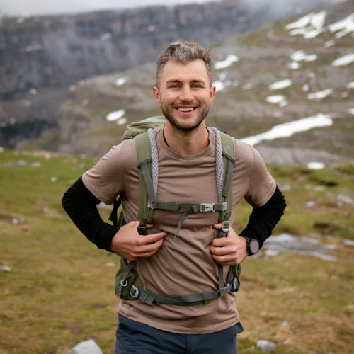 Uśmiechnięty mężczyzna z brodą stoi na świeżym powietrzu w górach, ubrany w plecak i odzież outdoorową obok Koszulka z wełny merino prosta damska mokka. Trawiasty, skalisty teren ze śnieżnymi plamami rozciąga się pod zachmurzonym niebem.