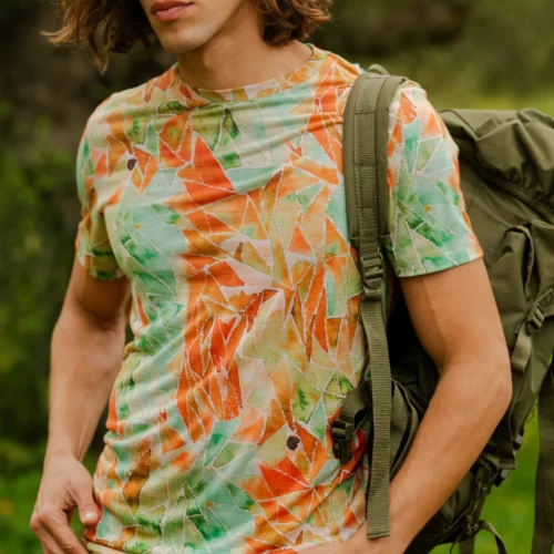 Mężczyzna z długimi brązowymi włosami, ubrany w czerwone okulary przeciwsłoneczne, koszulkę z wełny merynosów z rękawem 3/4 w kolorze mokka, beżowe szorty i zielony plecak, stoi na zewnątrz w otoczeniu bujnej zieleni.