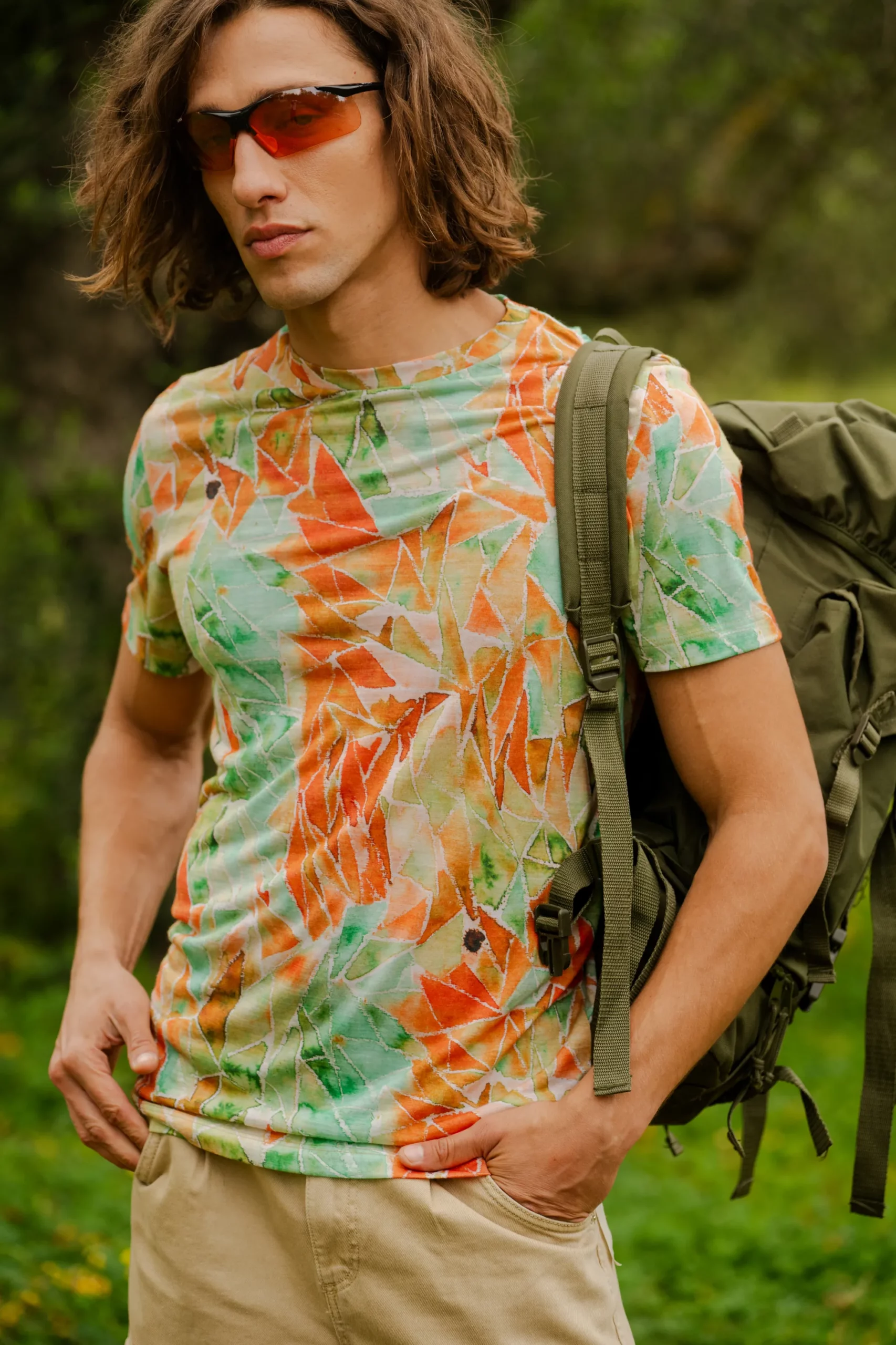 Mężczyzna z długimi brązowymi włosami, ubrany w czerwone okulary przeciwsłoneczne, koszulkę z wełny merynosów z rękawem 3/4 w kolorze mokka, beżowe szorty i zielony plecak, stoi na zewnątrz w otoczeniu bujnej zieleni.