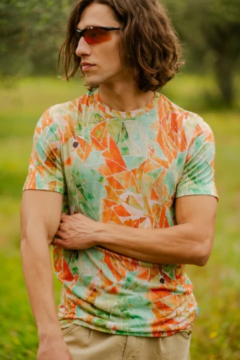 Mężczyzna z włosami do ramion i okularami przeciwsłonecznymi stoi na zewnątrz, z jedną ręką zgiętą w poprzek tułowia, ubrany w koszulkę z wełny merino z rękawem 3/4 mokka.