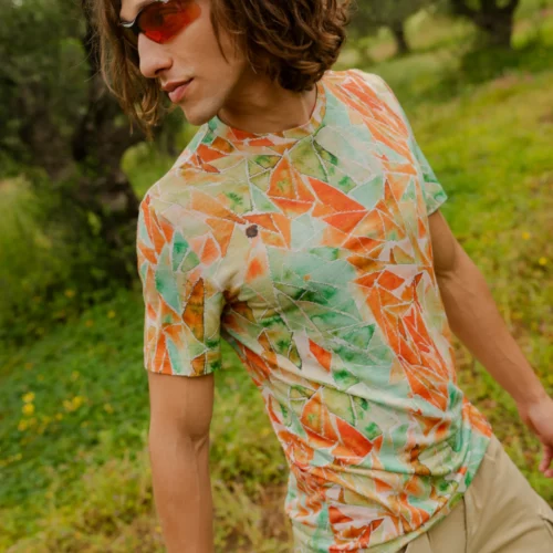 Mężczyzna z brązowymi włosami do ramion, pomarańczowymi okularami przeciwsłonecznymi i beżowymi szortami stoi na zewnątrz w koszulce z wełny merino z rękawem 3/4 mokka.