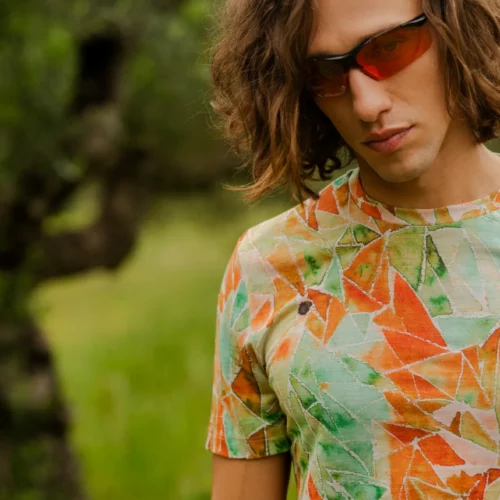 Osoba z brązowymi włosami do ramion i czerwonymi okularami przeciwsłonecznymi stoi na zewnątrz w rozmytej zieleni, ubrana w Koszulka z wełny merino z rękawem 3/4 mokka - kolorowy t-shirt z abstrakcyjnym pomarańczowym, zielonym i białym geometrycznym wzorem.