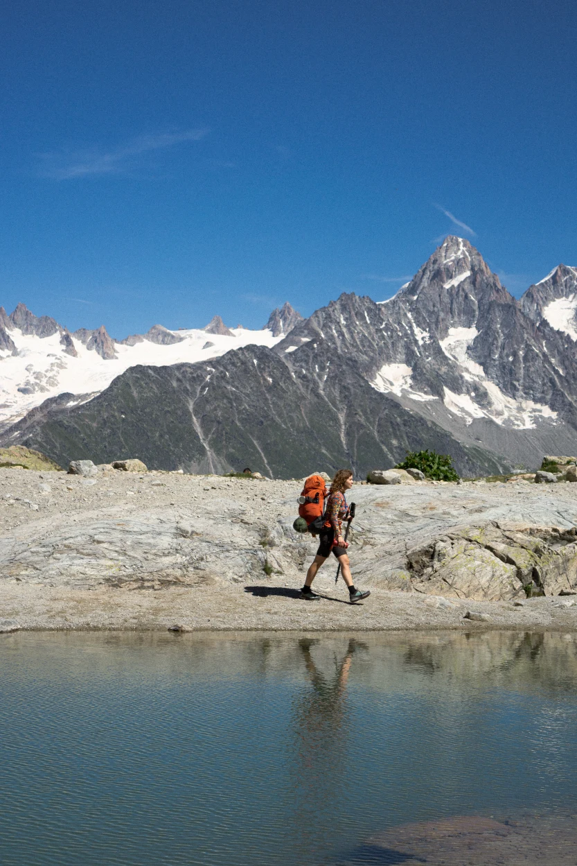 Turysta z pomarańczowym plecakiem spaceruje obok małego stawu, z ośnieżonymi górami i czystym, błękitnym niebem w tle.
