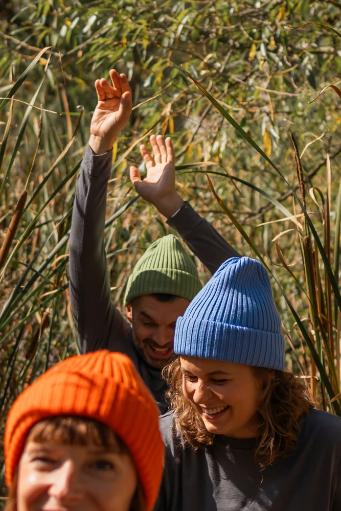 Trzy osoby w kolorowych dzianinowych czapkach - pomarańczowej, zielonej i niebieskiej - stoją wśród wysokiej trawy na zewnątrz. Są uśmiechnięci i radośni, a jedna osoba podnosi ręce w tle. Światło słoneczne tworzy ciepłą, radosną atmosferę.