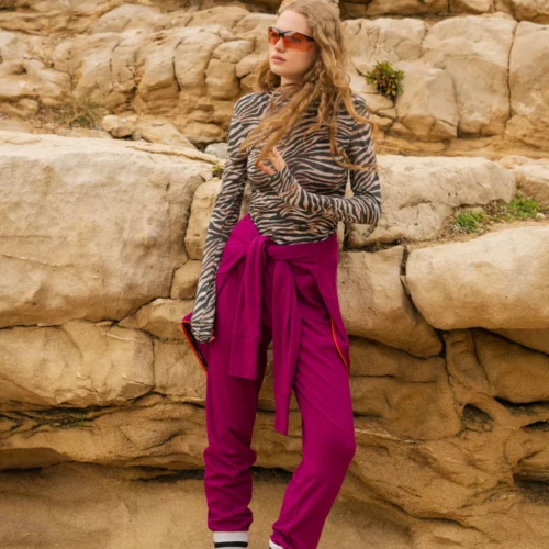 Osoba pozuje przed dużymi formacjami skalnymi, ubrana w okulary przeciwsłoneczne, bluzkę z długim rękawem w zebrę, karmazynowe spodnie z wysokim stanem i wiązaną talią, skarpetki w paski i grube trampki.