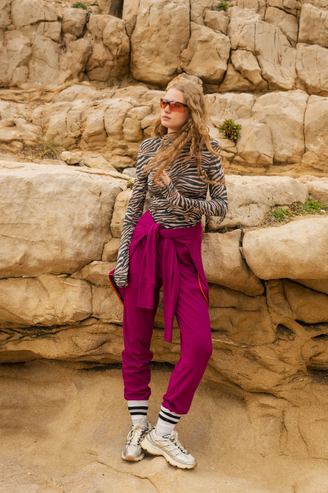Osoba pozuje przed dużymi formacjami skalnymi, ubrana w okulary przeciwsłoneczne, bluzkę z długim rękawem w zebrę, karmazynowe spodnie z wysokim stanem i wiązaną talią, skarpetki w paski i grube trampki.