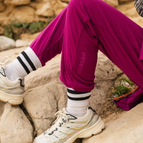 Osoba ubrana w karmazynowe joggery, białe buty sportowe i białe skarpetki w czarne paski siedzi na skalistym terenie. Widoczna jest tylko jej dolna część ciała i część fioletowej torby.