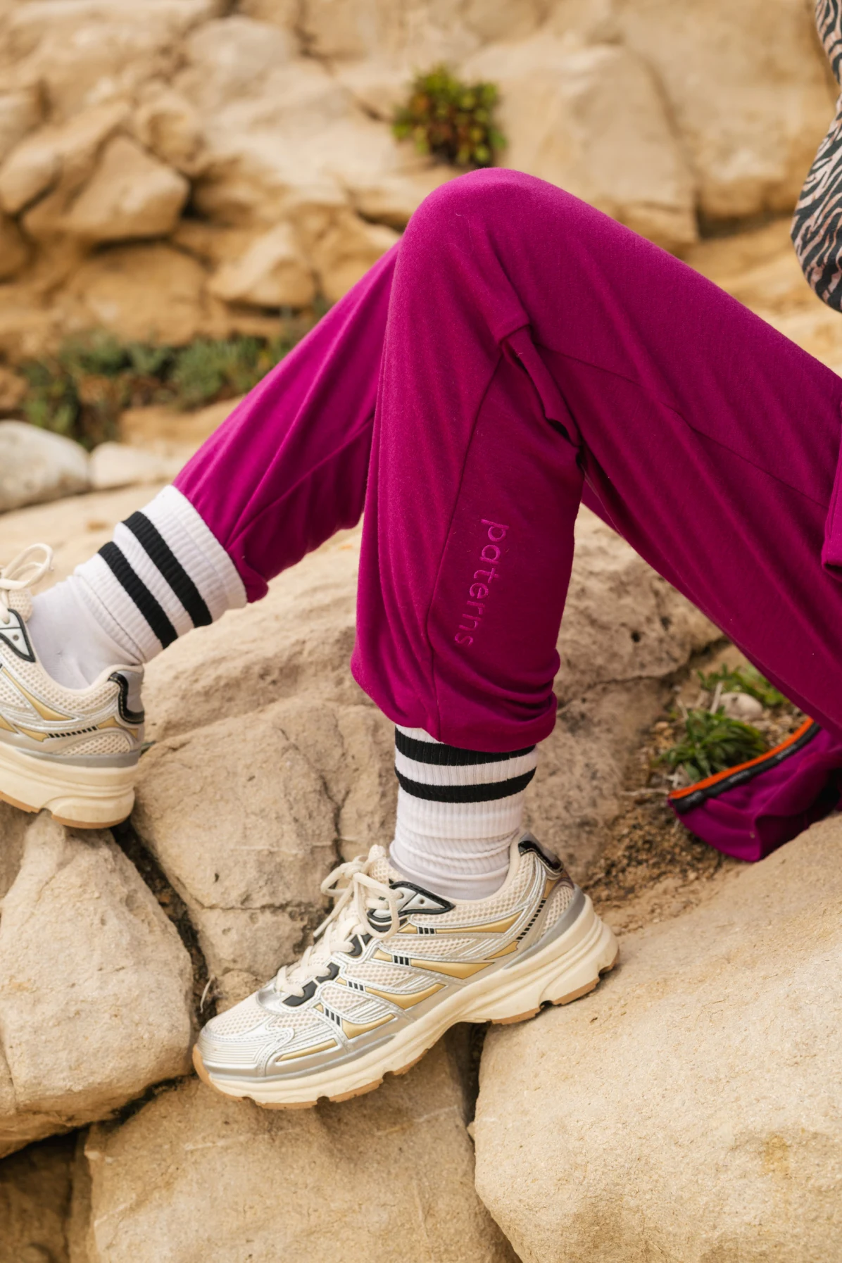 Osoba ubrana w karmazynowe joggery, białe buty sportowe i białe skarpetki w czarne paski siedzi na skalistym terenie. Widoczna jest tylko jej dolna część ciała i część fioletowej torby.
