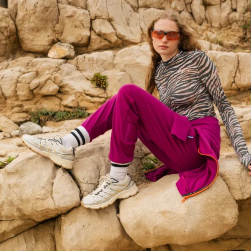 Osoba w bluzce z nadrukiem zebry, purpurowych spodniach, białych tenisówkach i pomarańczowych okularach przeciwsłonecznych siedzi na dużych skałach na zewnątrz, na skalistym tle.