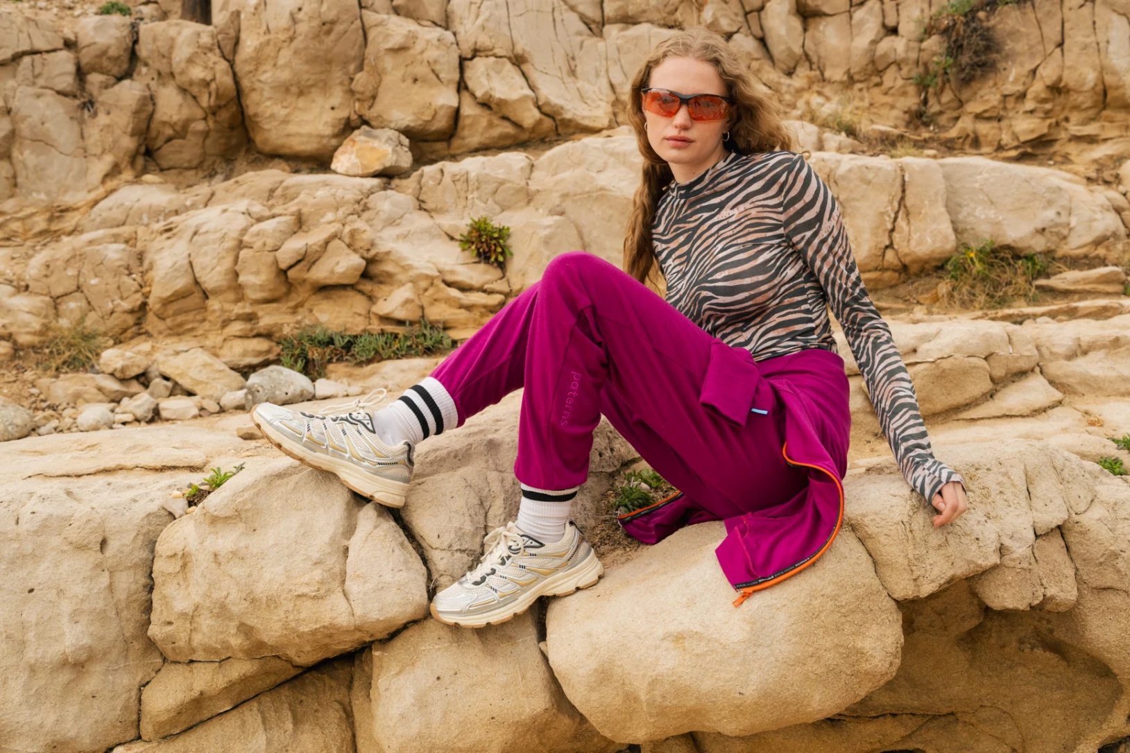 Osoba w bluzce z nadrukiem zebry, purpurowych spodniach, białych tenisówkach i pomarańczowych okularach przeciwsłonecznych siedzi na dużych skałach na zewnątrz, na skalistym tle.
