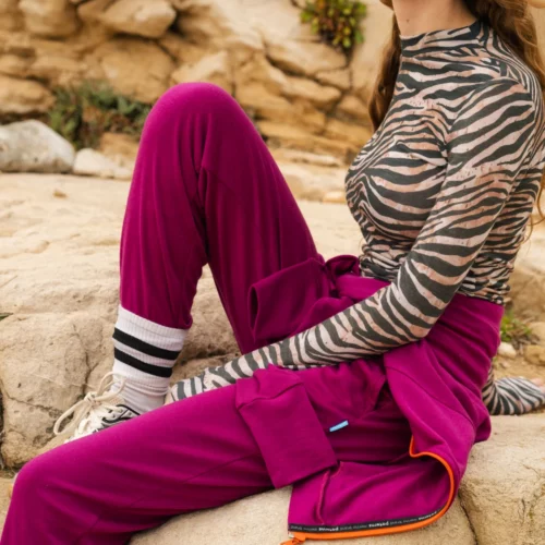 Kobieta z długimi falującymi włosami i pomarańczowymi okularami przeciwsłonecznymi siedzi na skale. Ma na sobie bluzkę w paski zebry, karmazynowe spodnie, pasującą kurtkę wokół talii, skarpetki w paski i trampki. Tło stanowi skalisty teren.