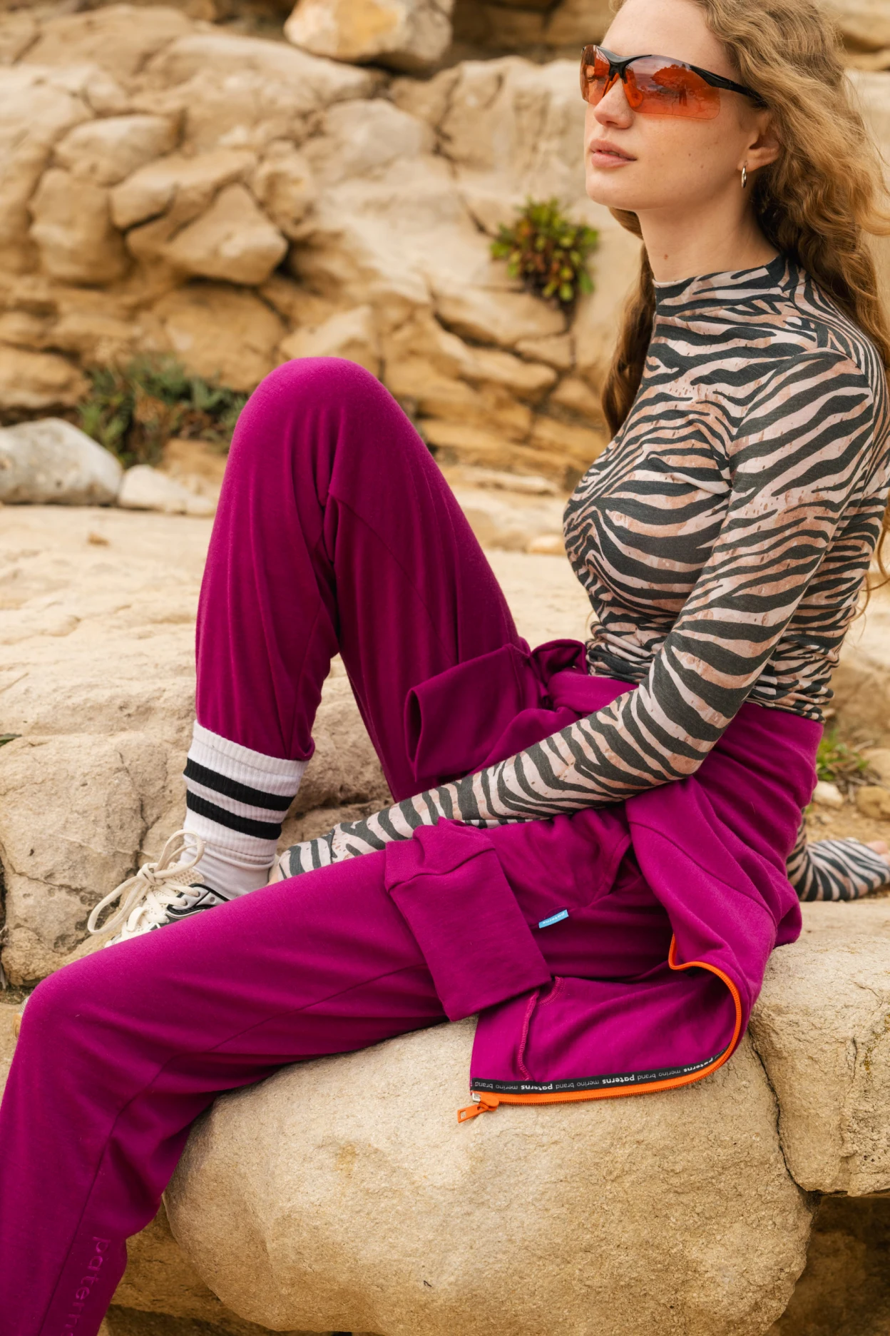 Kobieta z długimi falującymi włosami i pomarańczowymi okularami przeciwsłonecznymi siedzi na skale. Ma na sobie bluzkę w paski zebry, karmazynowe spodnie, pasującą kurtkę wokół talii, skarpetki w paski i trampki. Tło stanowi skalisty teren.