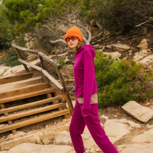 Osoba ubrana w jaskrawopomarańczową czapkę, okulary przeciwsłoneczne, karmazynowy dres i białe trampki spaceruje na zewnątrz w pobliżu drewnianych schodów i zieleni na kamienistej ścieżce.