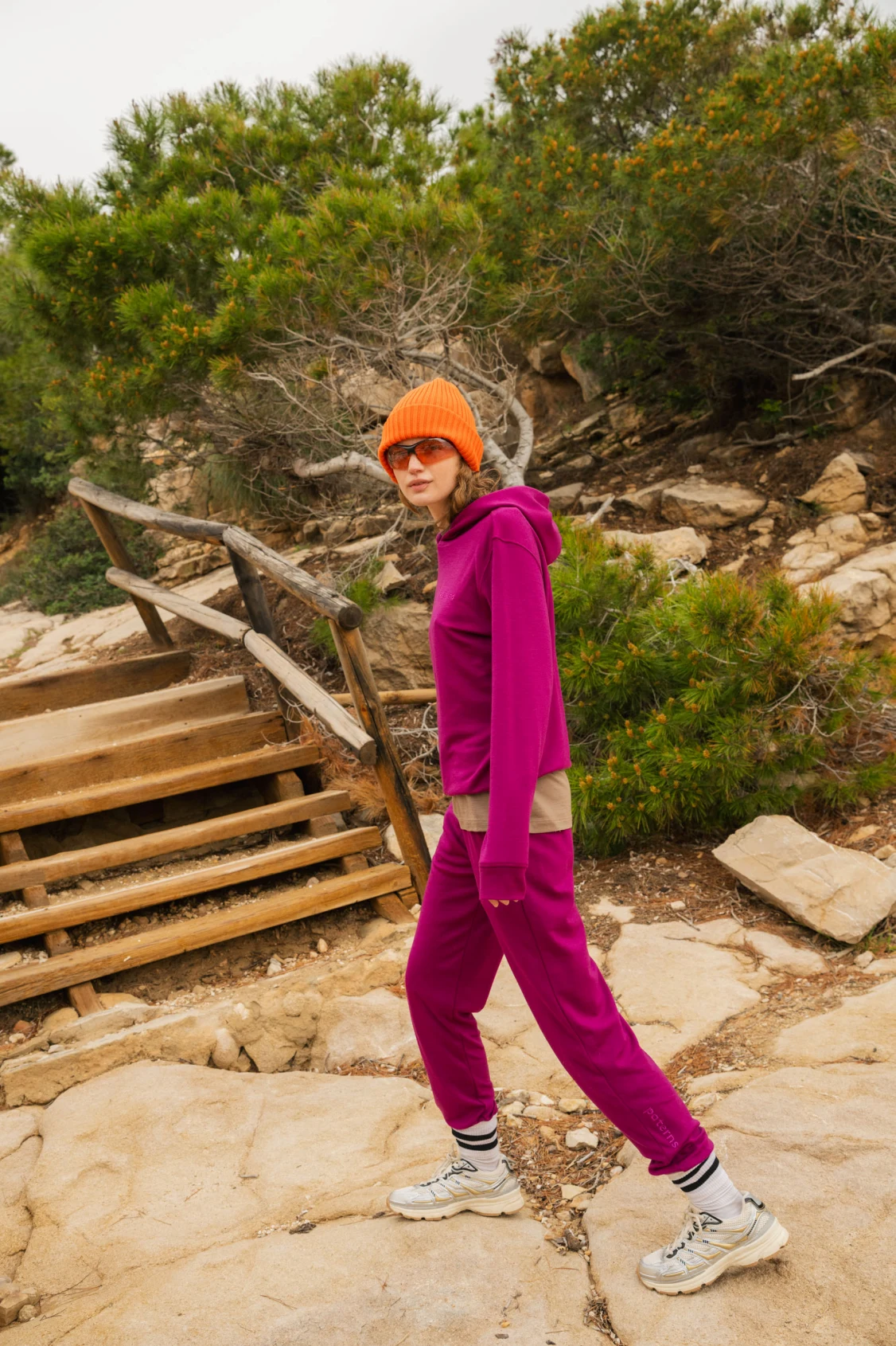 Osoba ubrana w jaskrawopomarańczową czapkę, okulary przeciwsłoneczne, karmazynowy dres i białe trampki spaceruje na zewnątrz w pobliżu drewnianych schodów i zieleni na kamienistej ścieżce.