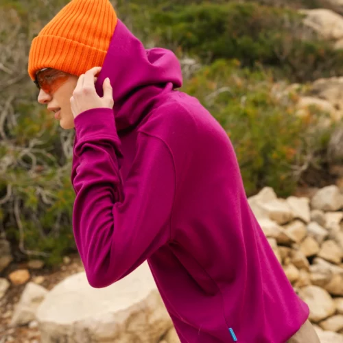Osoba na zewnątrz ubrana w jasnopomarańczową czapkę, okulary przeciwsłoneczne i magentową bluzę z kapturem. W tle widoczny jest skalisty teren i zielone krzewy.