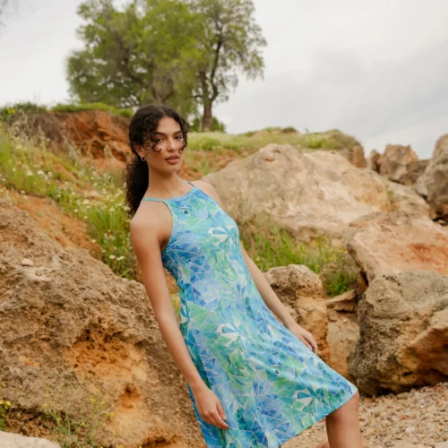 Kobieta z kręconymi włosami patrzy w kamerę na zewnątrz, stojąc na skalistym terenie z zielenią i dużymi kamieniami, ubrana w koszulkę z wełny merino z rękawem 3/4 mokka.