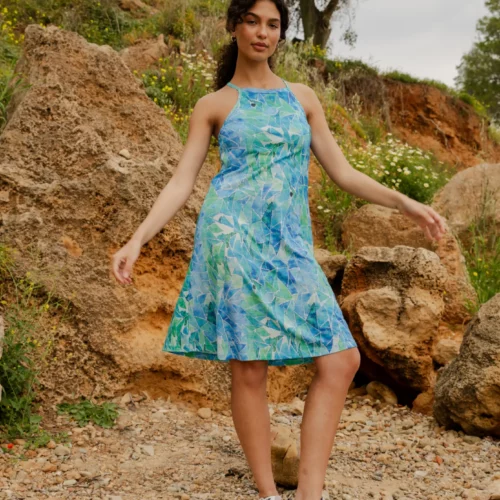 Kobieta ubrana w koszulkę z wełny merino z rękawem 3/4 mokka stoi na zewnątrz na skalistym podłożu, otoczona dużymi skałami i zielenią, pozując z lekko uniesioną ręką.