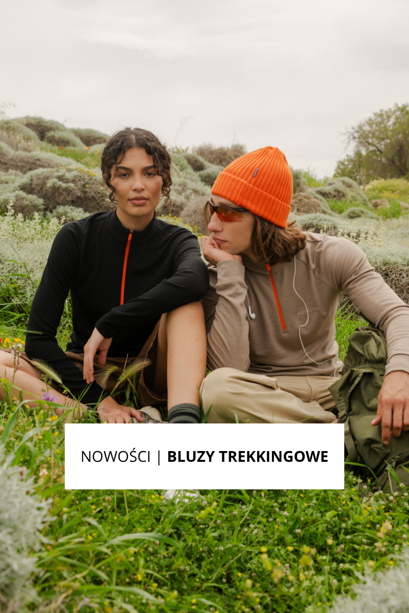 Dwoje młodych dorosłych siedzi na zewnątrz na trawiastym terenie. Kobieta ma na sobie czarną kurtkę i szorty, a mężczyzna beżową kurtkę, pomarańczową czapkę i okulary przeciwsłoneczne. Oboje wyglądają na zrelaksowanych. Na banerze widnieje napis: NOWOŚCI | BLUZY TREKKINGOWE.