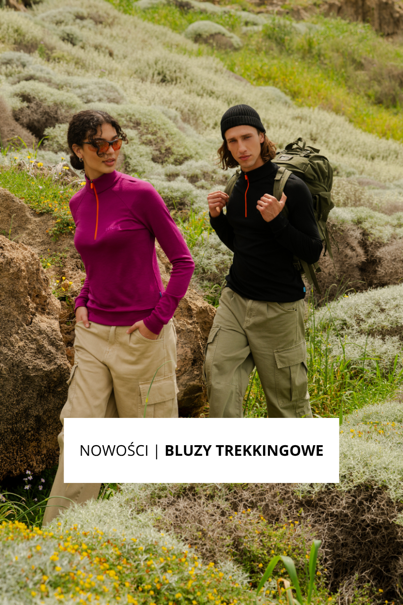 Dwie osoby wędrujące po trawiastym, skalistym krajobrazie. Jedna z nich ma na sobie magentową bluzę trekkingową i okulary przeciwsłoneczne, a druga czarną bluzę trekkingową, czarną czapkę i plecak. Tekst: Nowości | Bluzy Trekkingowe.