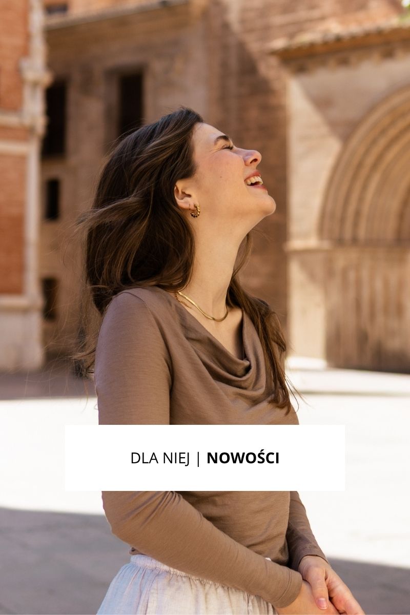 Uśmiechnięta kobieta z długimi brązowymi włosami, ubrana w jasnobrązowy top i złote kolczyki, stoi na zewnątrz w pobliżu zabytkowych kamiennych budynków. Nakładka z tekstem w języku polskim: Dla Niej | Nowości.