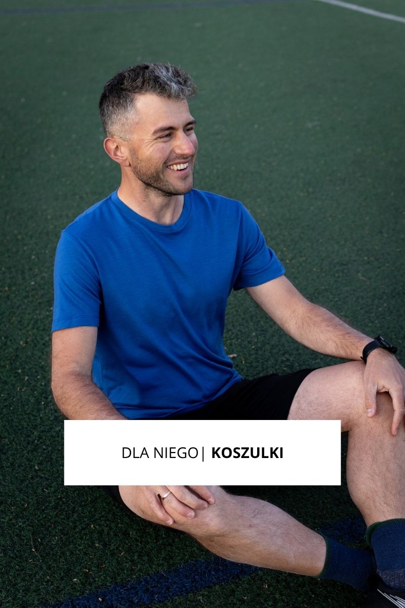 Mężczyzna z krótkimi włosami i brodą, ubrany w niebieską koszulkę i czarne szorty, siedzi na boisku sportowym i uśmiecha się. Nałożony tekst brzmi DLA NIEGO | KOSZULKI.