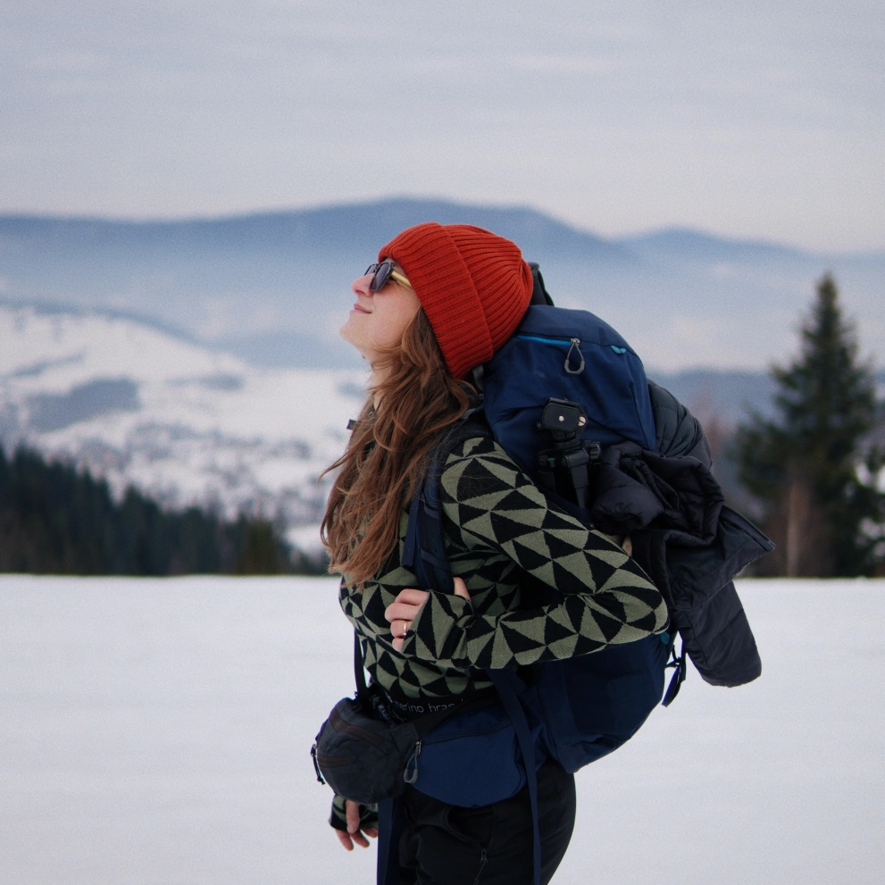 Kobieta w czerwonej czapce, okularach przeciwsłonecznych i swetrze w geometryczne wzory stoi w śnieżnym krajobrazie z dużym plecakiem turystycznym, patrząc w górę. W tle widoczne są ośnieżone góry i drzewa.