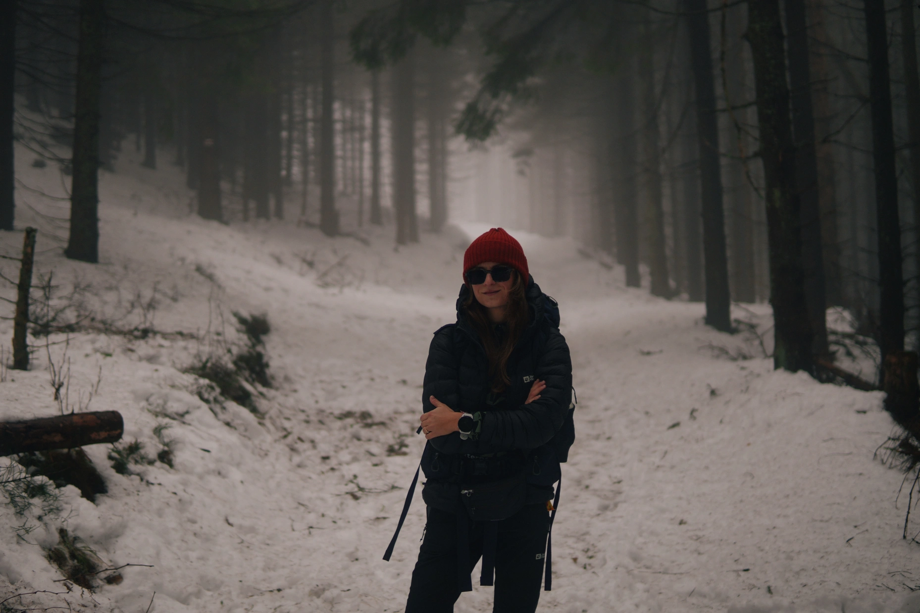 Osoba ubrana w czerwoną czapkę, ciemne okulary przeciwsłoneczne i czarną kurtkę stoi na pokrytej śniegiem ścieżce w mglistym, gęstym lesie. Drzewa są wysokie, a atmosfera jest mglista i spokojna.