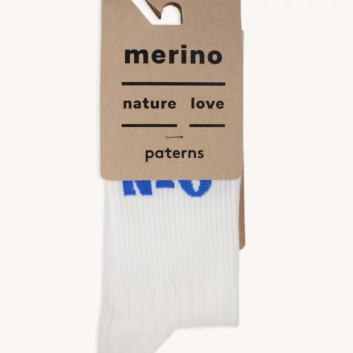 Para białych Skarpety trekkingowe merino z napisem RÓB NIC w kolorze czarnym, zapakowane na brązowym kartonie z napisem "nature love patterns". Niebieskie litery są częściowo widoczne na skarpetach.