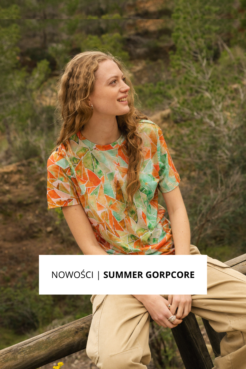 Osoba z długimi falującymi włosami siedzi na zewnątrz na drewnianym płocie, ubrana w kolorową koszulkę z geometrycznymi wzorami i jasne spodnie. W tle drzewa i skalisty teren. Tekst brzmi: NOWOŚCI | LETNI GORPCORE.