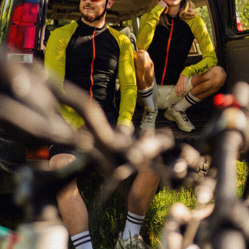 Dwie rowerzystki ubrane w identyczne czarne koszulki rowerowe z wełny merynosowej z długim rękawem dla kobiet, relaksują się i uśmiechają na tylnym siedzeniu SUV-a, a na słonecznym pierwszym planie widać rozmyte sylwetki rowerów.