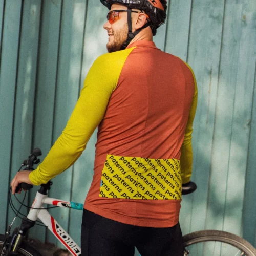 Rowerzysta w kasku, okularach przeciwsłonecznych i męskiej koszulce rowerowej z wełny merynosowej w kolorze ceglastym, z długimi rękawami, stoi obok białego roweru, odwrócony plecami do turkusowego drewnianego ogrodzenia.