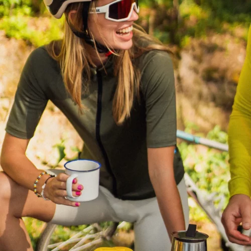 Uśmiechnięta kobieta w stroju rowerowym i kasku, ubrana w „Koszulkę rowerową z wełny merynosowej z krótkim rękawem dla kobiet w kolorze oliwkowym”, trzyma biały kubek, siedząc na świeżym powietrzu obok roweru, a na stole przed nią stoją dzbanki z kawą i żółty termos.