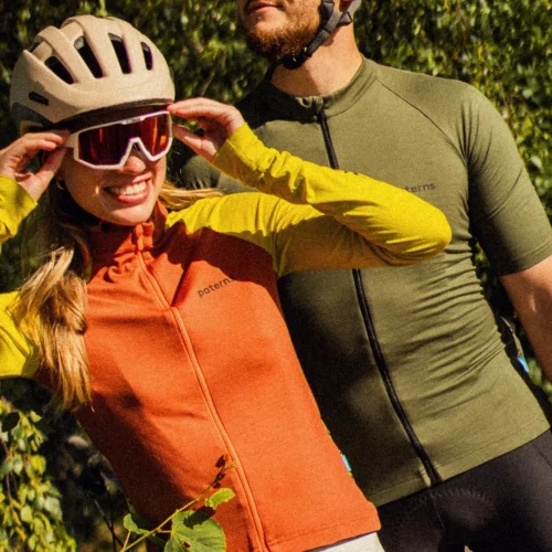 Dwoje rowerzystów w kaskach i okularach przeciwsłonecznych stoi na świeżym powietrzu. Kobieta z przodu ma na sobie pomarańczowo-żółtą koszulkę i uśmiecha się, poprawiając kask. Za nią stoi mężczyzna, wpatrujący się w dal, ubrany w męską koszulkę rowerową z wełny merynosowej z krótkim rękawem w kolorze oliwkowym.