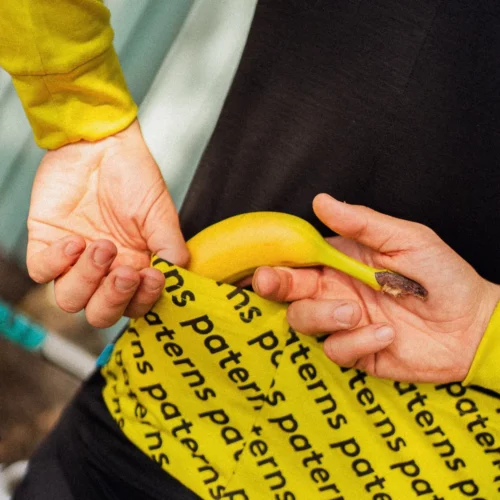Osoba ubrana w czarną męską koszulkę rowerową z długim rękawem z wełny merynosowej wkłada banana do tylnej kieszeni swojej koszulki rowerowej.