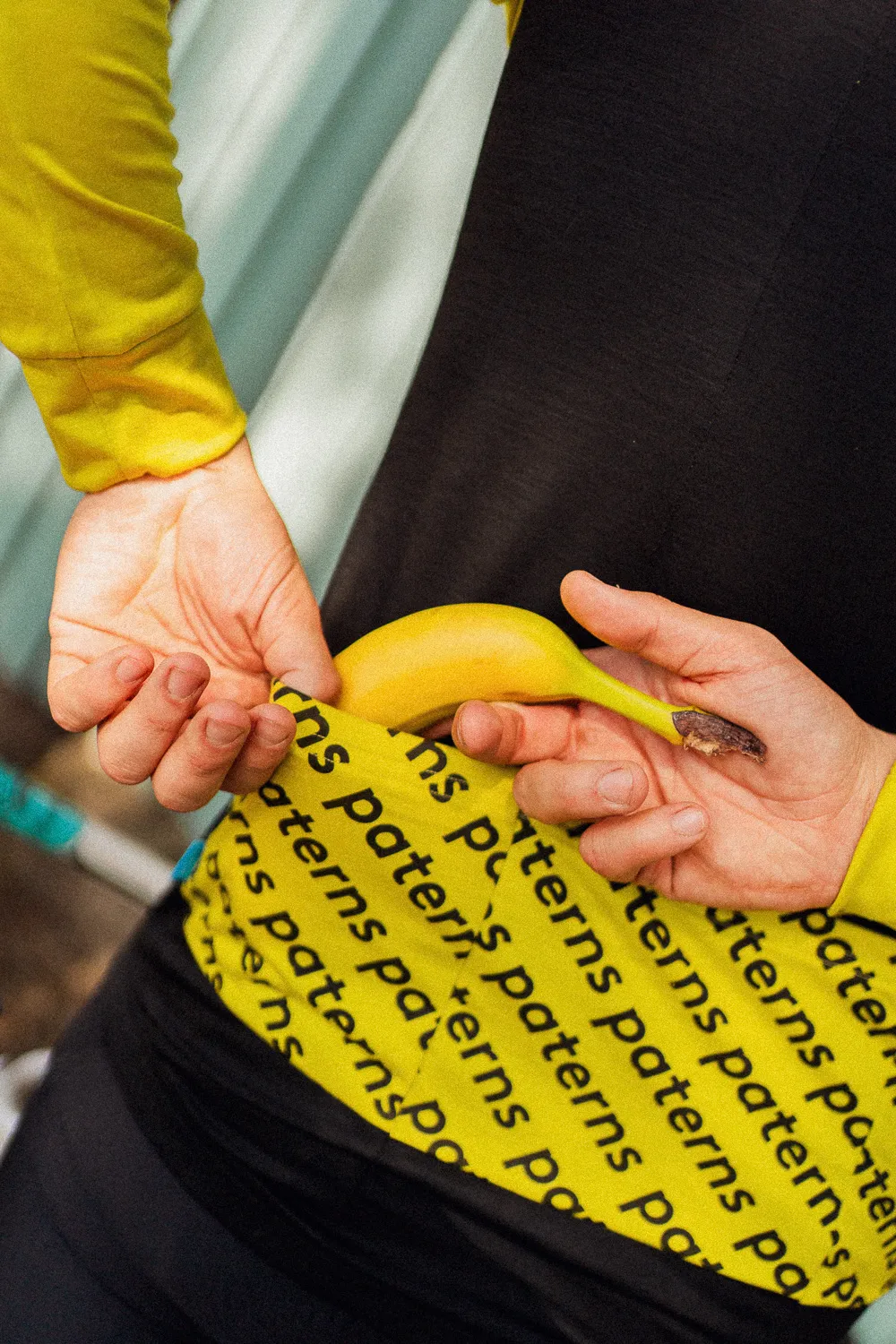 Osoba ubrana w czarną męską koszulkę rowerową z długim rękawem z wełny merynosowej wkłada banana do tylnej kieszeni swojej koszulki rowerowej.