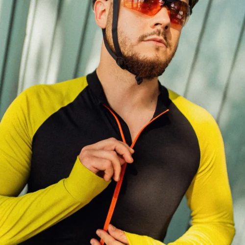 Mężczyzna w różowym kasku, czerwonych sportowych okularach przeciwsłonecznych i „czarnej męskiej koszulce rowerowej z długim rękawem z wełny merynosowej” zapina zamek błyskawiczny na swojej koszulce rowerowej, przebywając na świeżym powietrzu w słońcu.