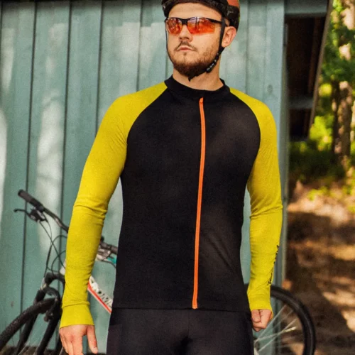 Mężczyzna w okularach przeciwsłonecznych i czarnej koszulce rowerowej z wełny merynosowej z żółtymi rękawami (Koszulka rowerowa z wełny merynosowej z długim rękawem, męska, czarna) stoi przy szopie wraz ze swoim rowerem, a w tle promienie słońca prześwitują przez drzewa.