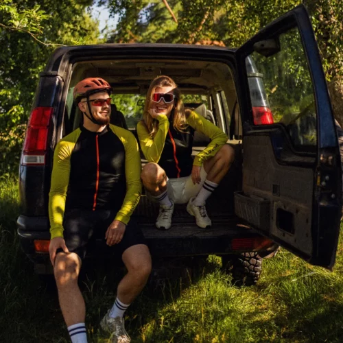 Dwóch rowerzystów odpoczywa w bagażniku SUV-a w skąpanym w słońcu lesie, uśmiechając się. Obaj mają na sobie identyczne kaski, okulary przeciwsłoneczne oraz czarną męską koszulkę rowerową z wełny merynosowej z długim rękawem: Koszulka rowerowa z wełny merynosowej z długim rękawem, męska, czarna.